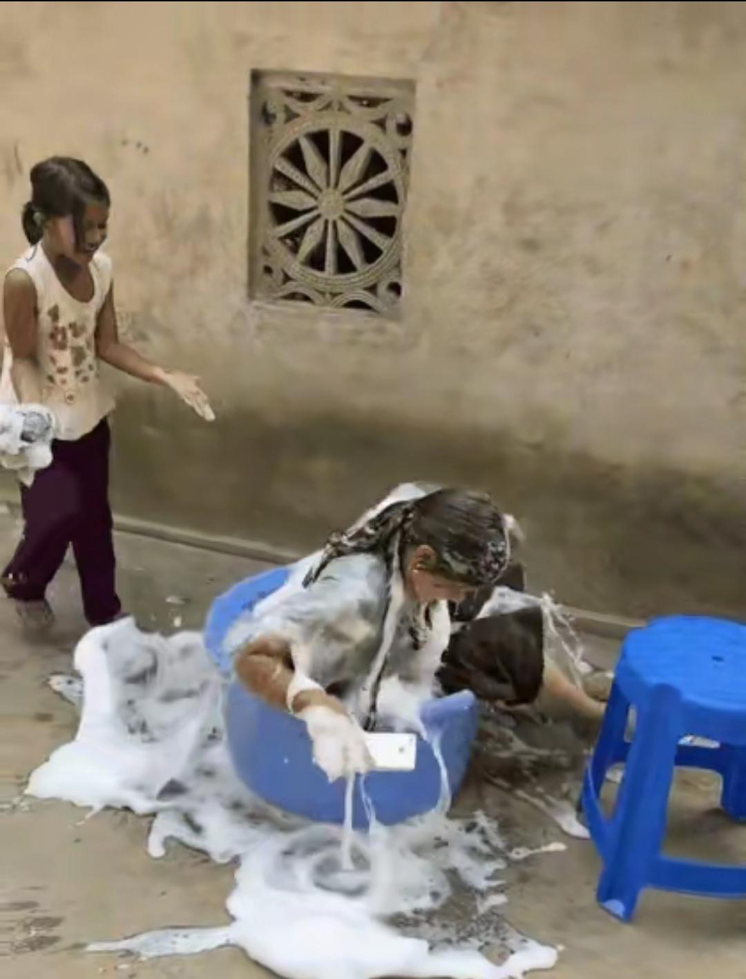 一个7岁左右的小孩坐在凳子上洗衣服。妈妈拿个手机打电话。也坐在女儿后边坐一个凳子