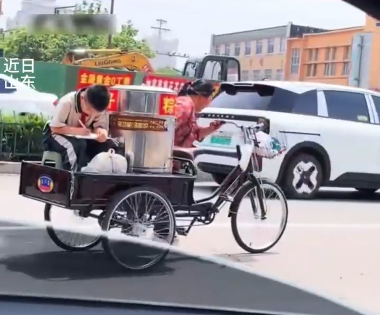 近日，在山东 一位妈妈蹬着装满粽子的小三轮车载着一男孩，男孩在颠簸中认真地写着作