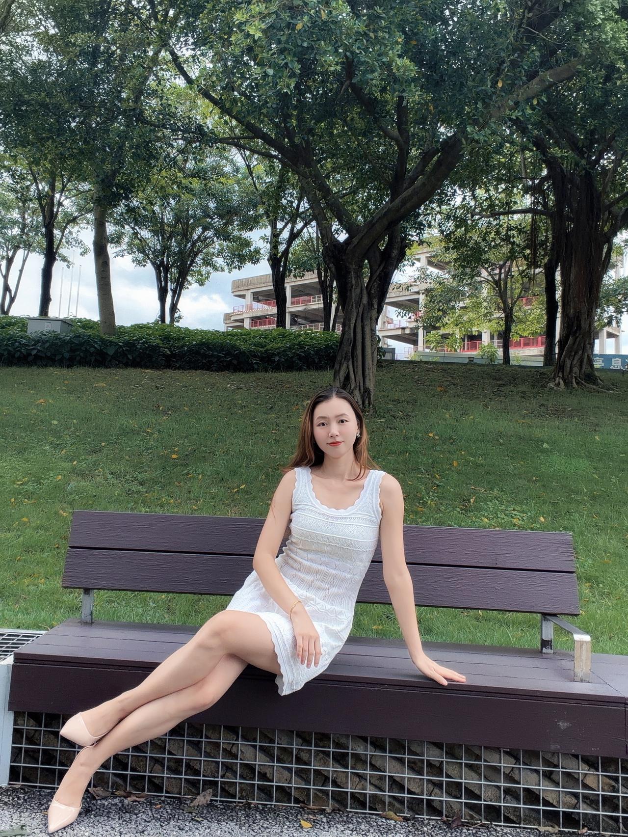 一个女孩坐在公园长椅上，背景是绿树和建筑。