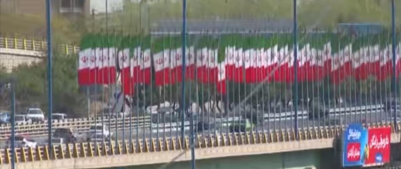 德黑兰街头悬挂着伊朗国旗，车水马龙的高速路与标志性的自由塔勾勒出城市风貌，与此同