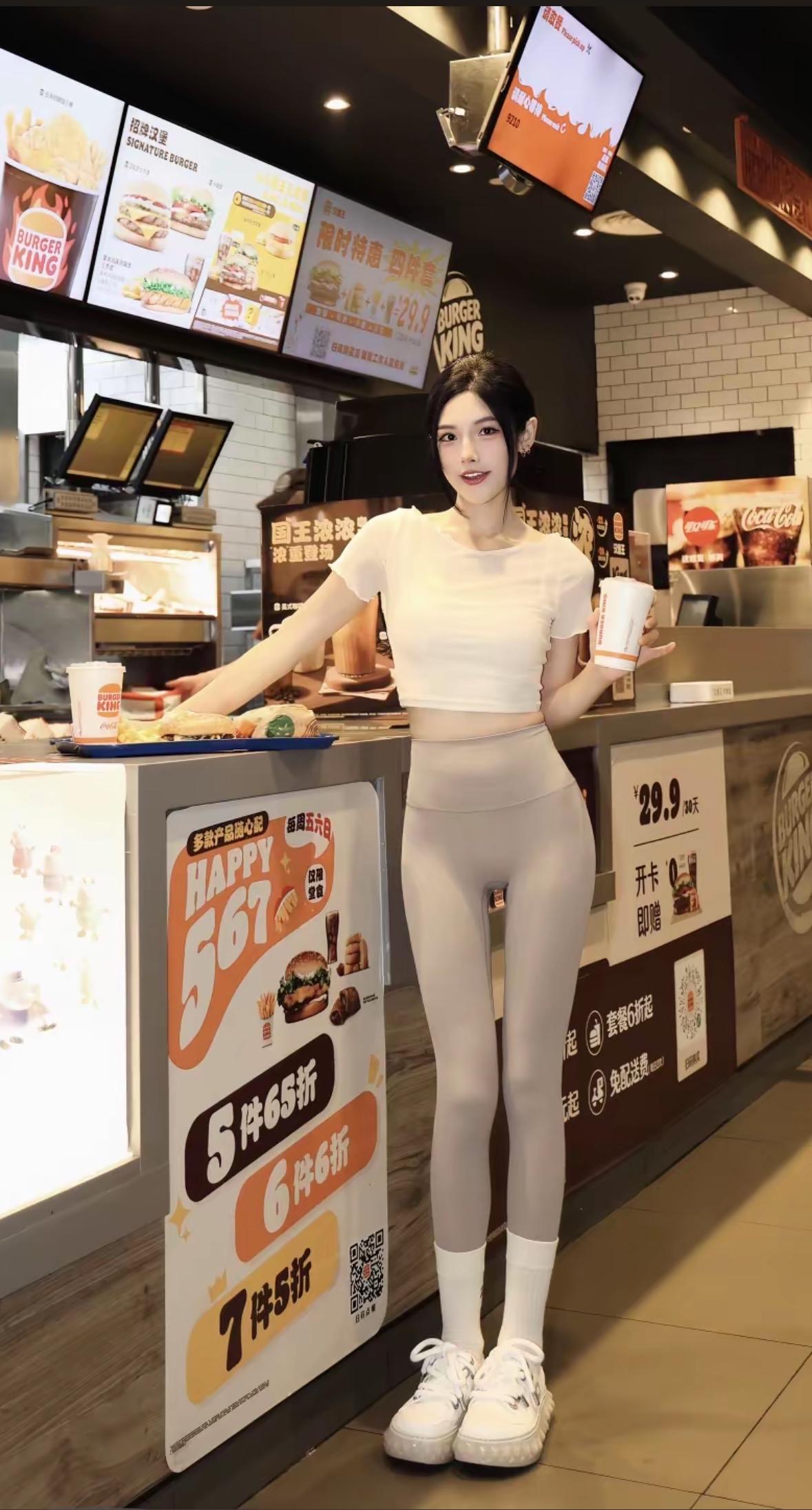 在汉堡王餐厅内，一位身穿白色短款上衣和浅灰色紧身裤的女生手持饮料杯，倚靠在点餐柜