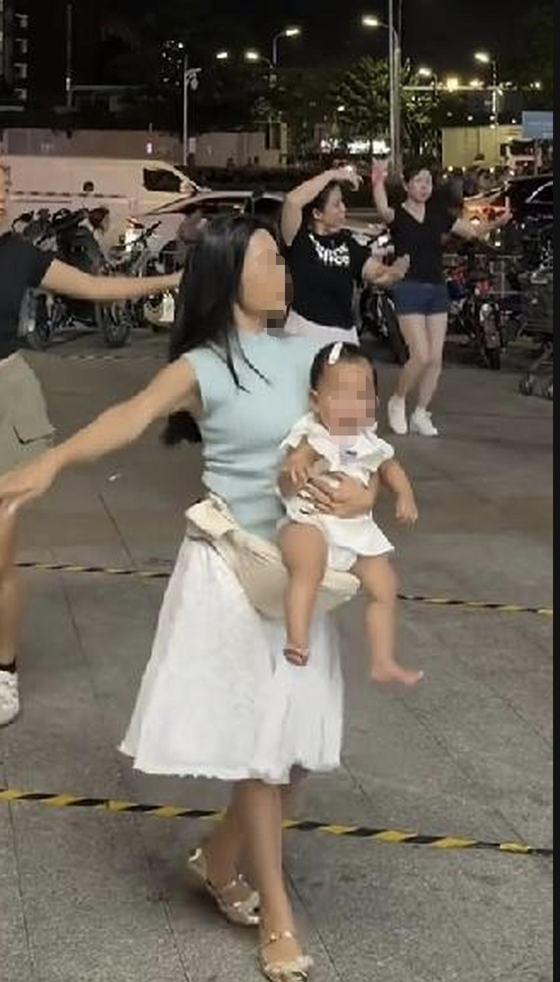 夜里广场舞热闹的场地，一名女子怀里抱着年幼的宝宝，跟着音乐跳广场舞。孩子明显状态