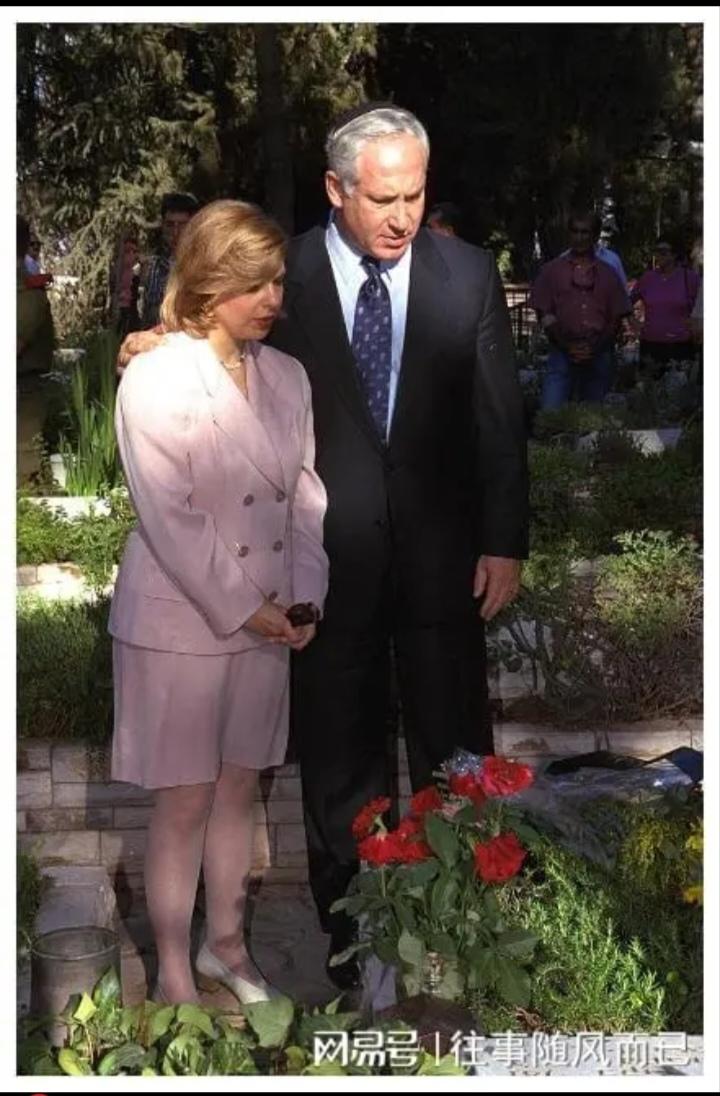 内塔尼亚胡的妻子一身粉色打扮，低头站在花草间，此刻的她有几分女人味。