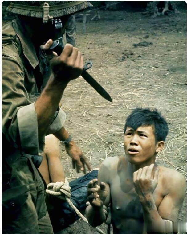 越南战争期间，一名美军搜索游击队下落时，手持军刀逼问当地村民的画面。
照片中男子