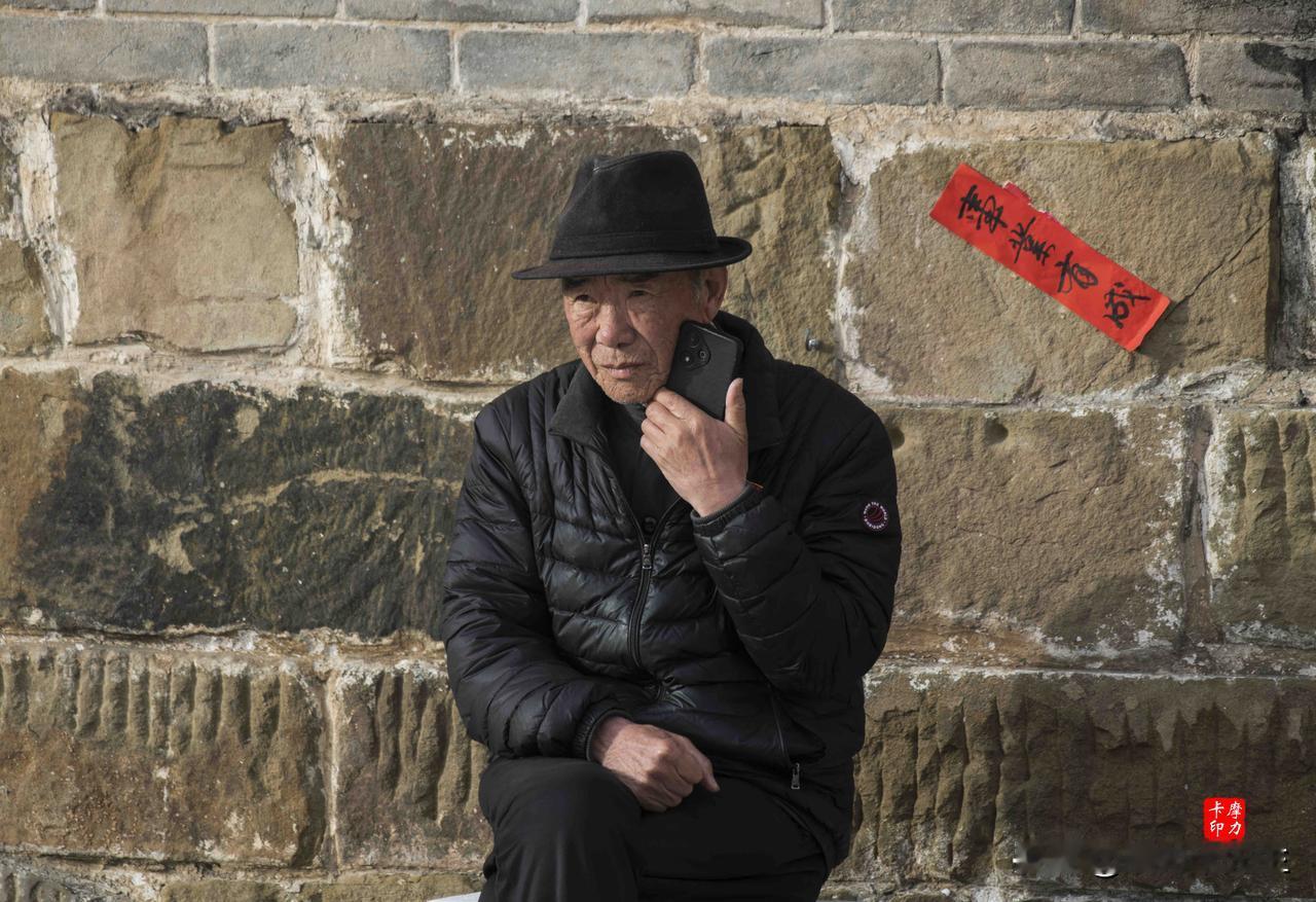 老墙根下，他戴着旧礼帽，对着手机轻声说着什么。身后红纸上“事业有成”的字，被风晒