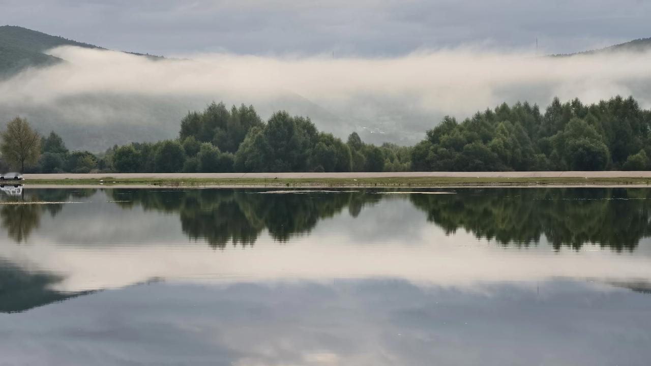 大兴安岭，晨雾缭绕，静谧湖面倒映着远山与绿树，宛如世外桃源。