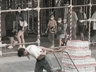 男士在街头拉着一车矿泉水吃力地行走，旁边逛街男士看到后，把手中的饮品递给自己女儿