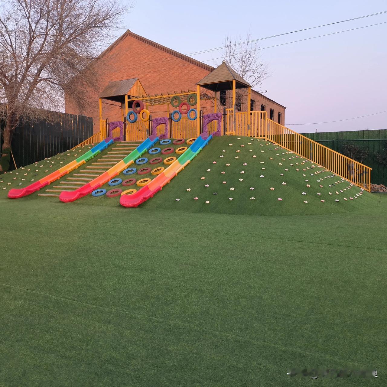 路过发现个超有意思的儿童游乐区！绿油油的仿真草坡上立着三条彩虹滑梯，红橙黄绿蓝的