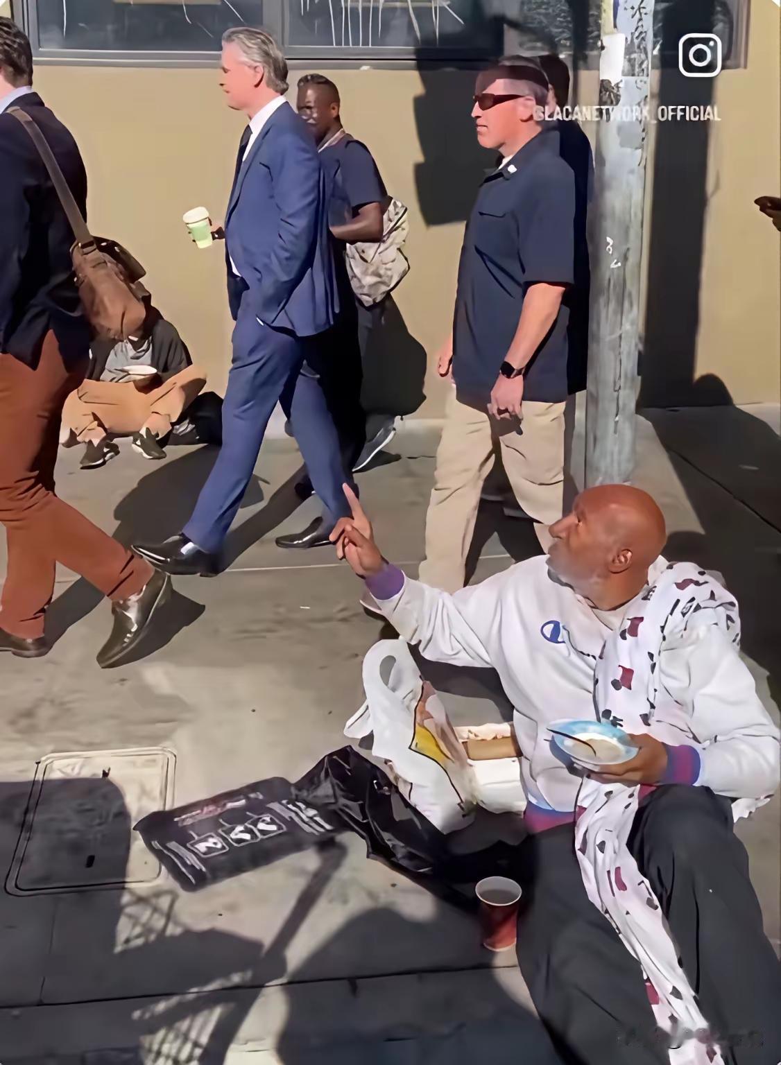 旁边保镖一刻也不敢松懈

美国加州州长纽森衣冠楚楚，单手插兜，手拿咖啡从流浪汉环