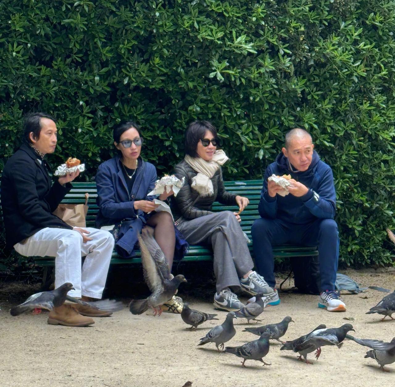 刘嘉玲、梁朝伟夫妻二人和友人一起看画展。梁朝伟光头造型坐在长椅上吃汉堡，好接地气