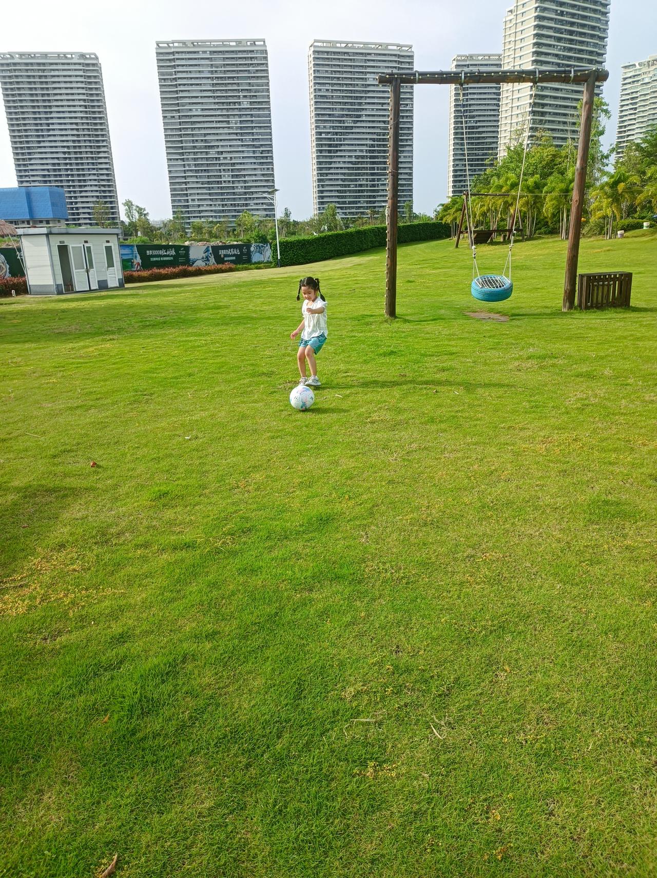 晒图笔记大赛 阳光洒满的公园，小女孩快乐地踢着足球，背景是现代化的高楼大厦。