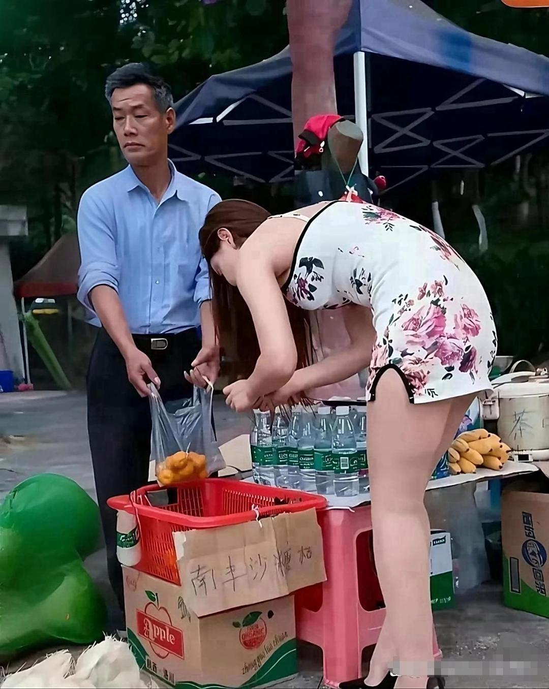 一个身穿花色超短旗袍，身材高挑的女孩躬着身子，正在全神贯注的挑选橘子！卖橘子的大
