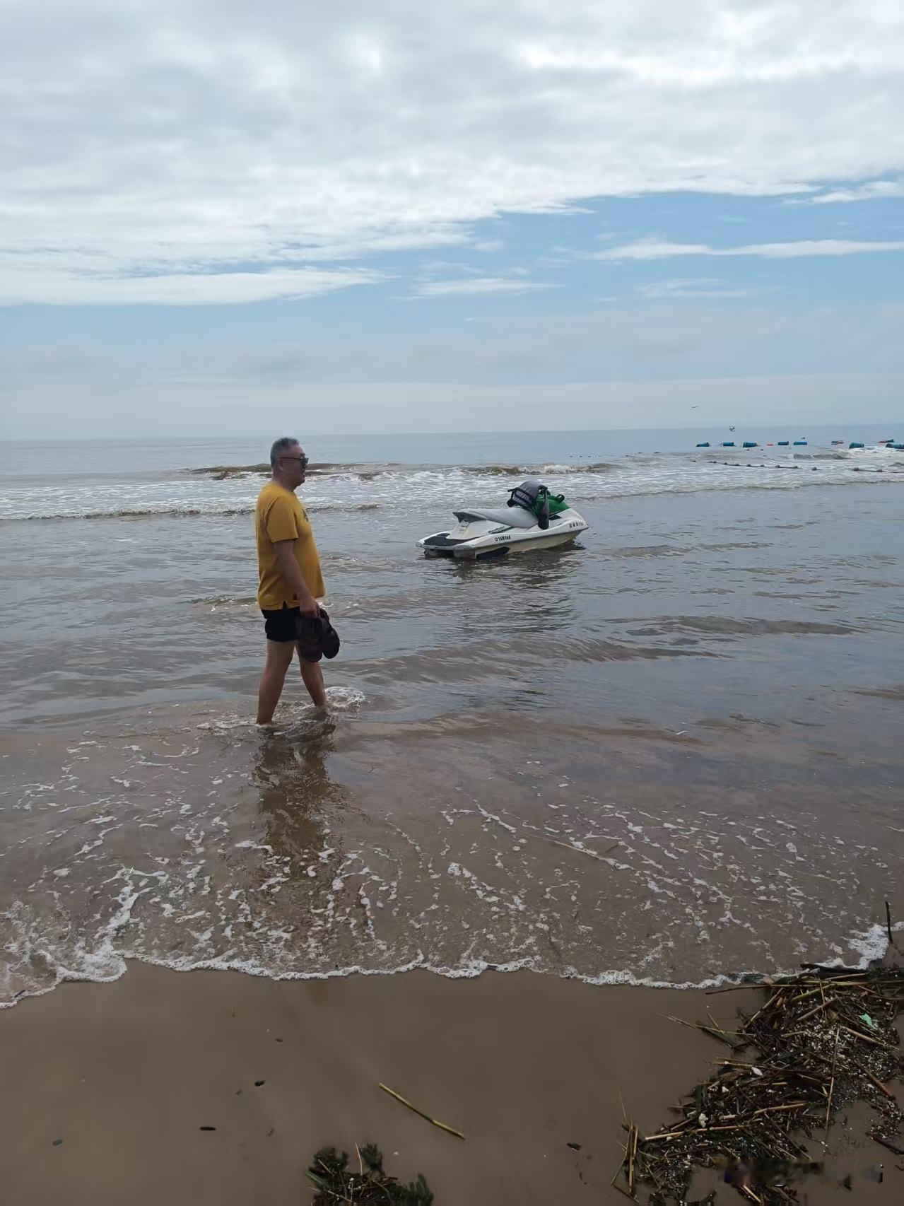 海边随性拍下这一幕：轻柔海浪漫上沙滩，浅海停着摩托艇，大叔赤脚蹚水慢行。云层舒展