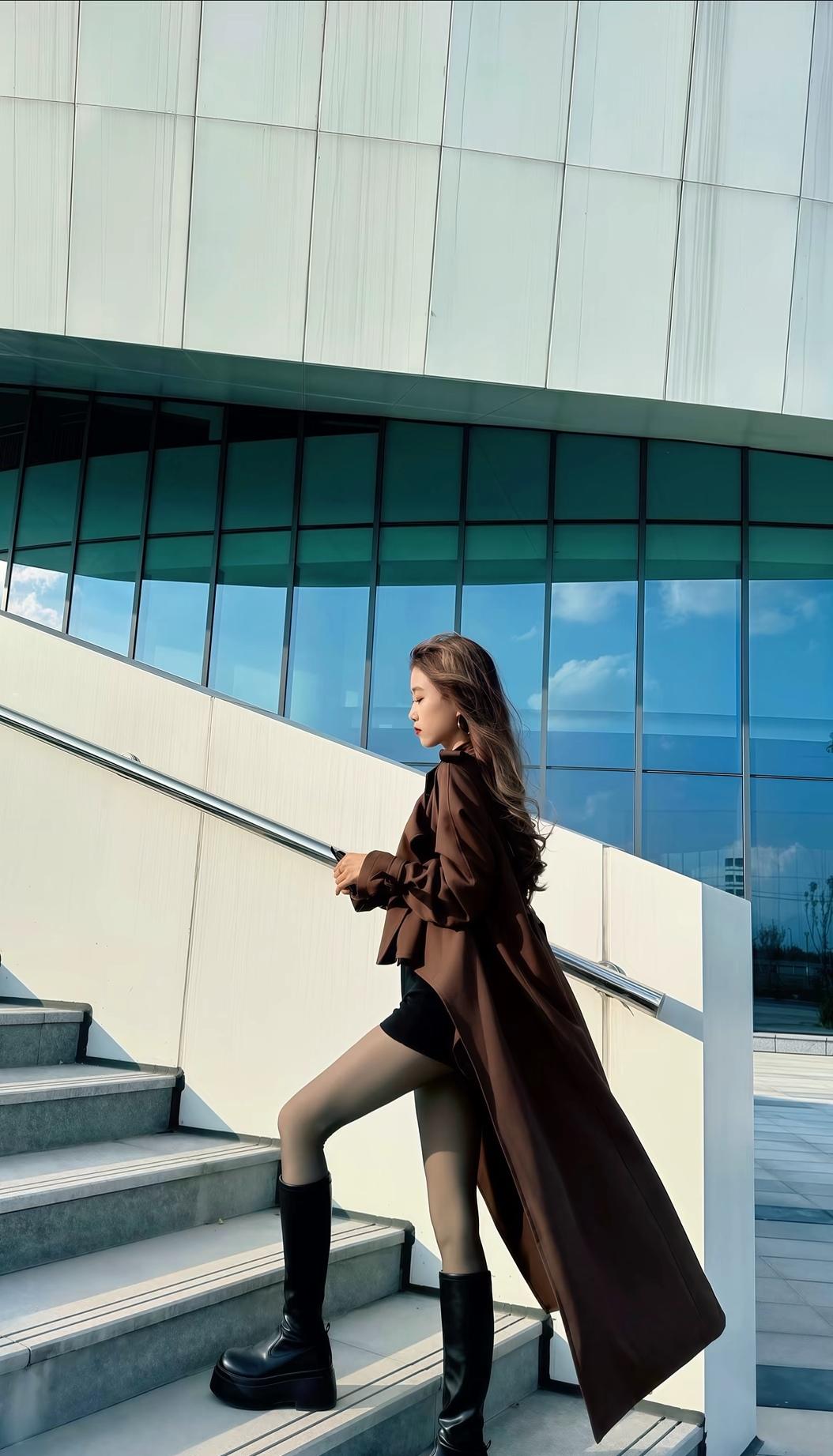 都市时尚风，棕色大衣搭配黑色靴子，走在现代建筑旁，尽显优雅与自信。