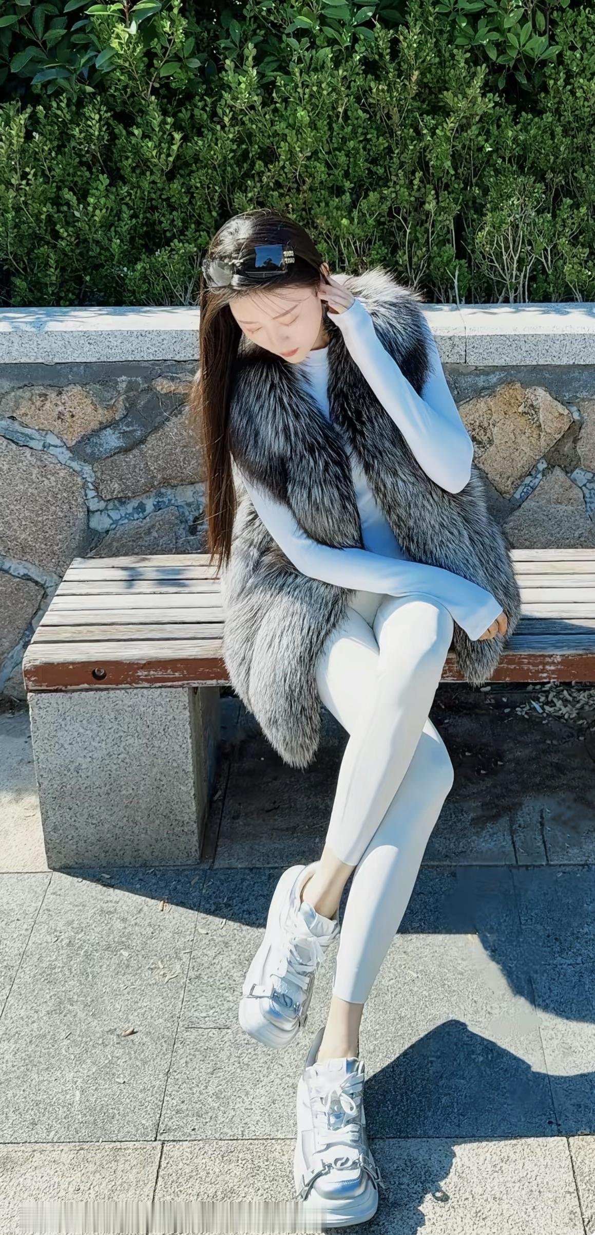 阳光明媚的午后，女孩静坐长椅。灰黑皮草马甲搭配白色紧身衣，脚踩厚底白鞋，慵懒又时