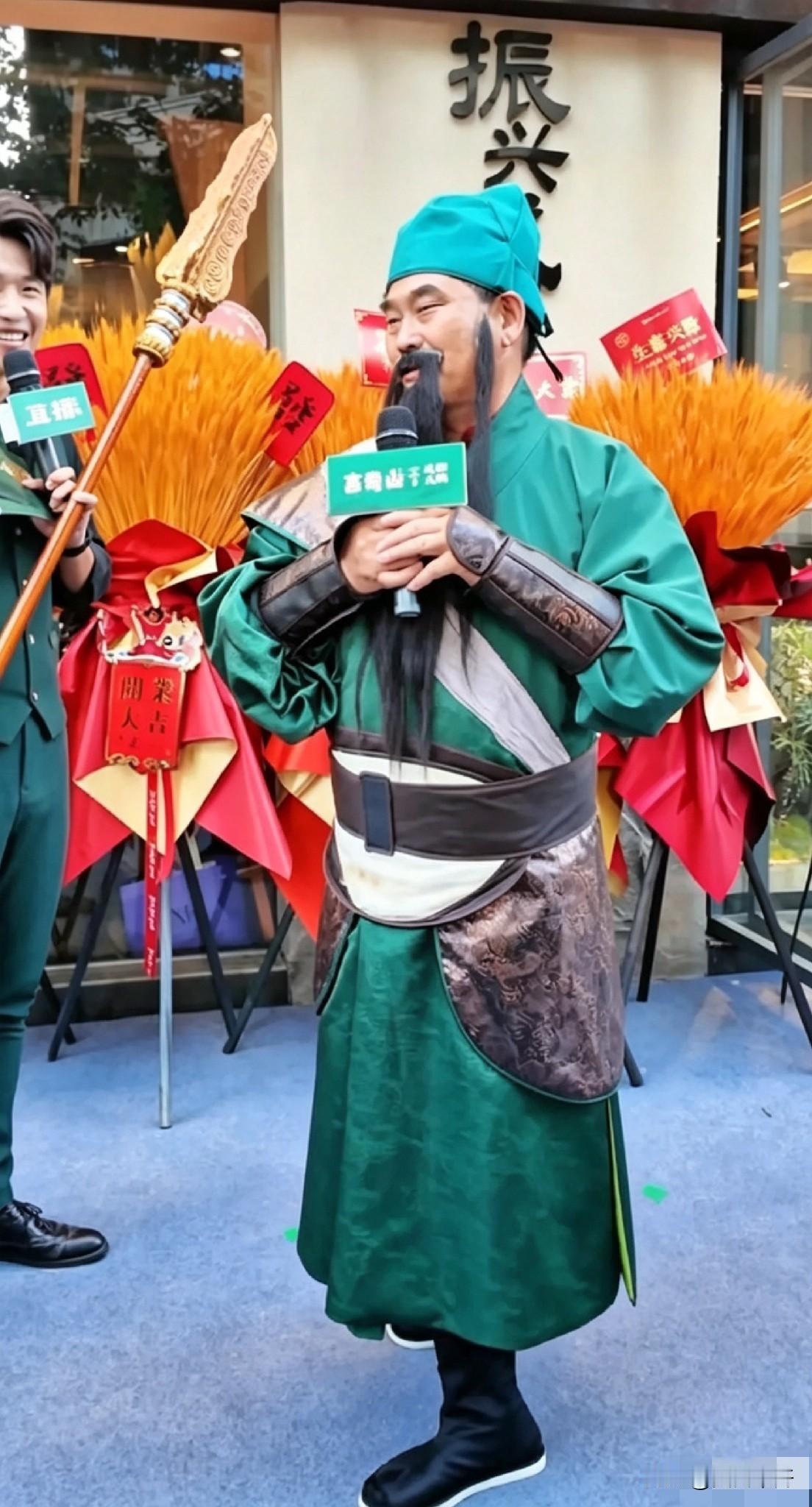 商演现场，大衣哥一身关公造型登场，绿袍长髯扮相十足，双手捧着话筒致辞，身后摆满开