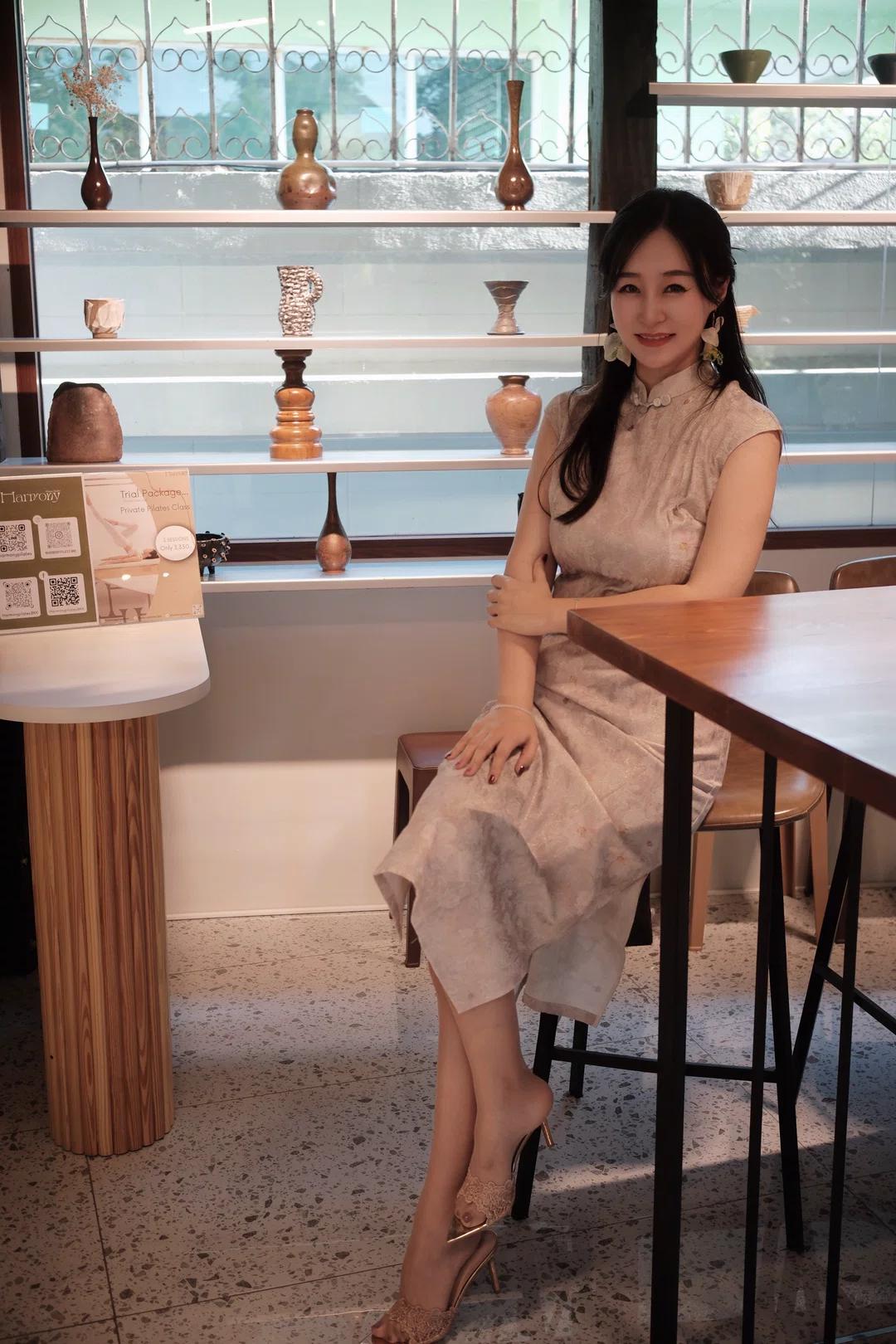 图片中是一位坐在木质长椅上的女性，她身穿浅色带有花纹的无袖旗袍，脚踩一双浅色高跟