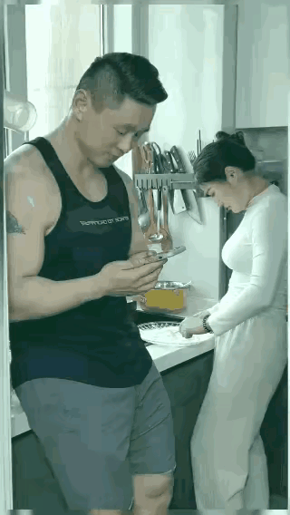 妻子在厨房和面做饭，丈夫在一旁看着手机傻笑，还对着手机里的美女狠狠的亲了两口。这