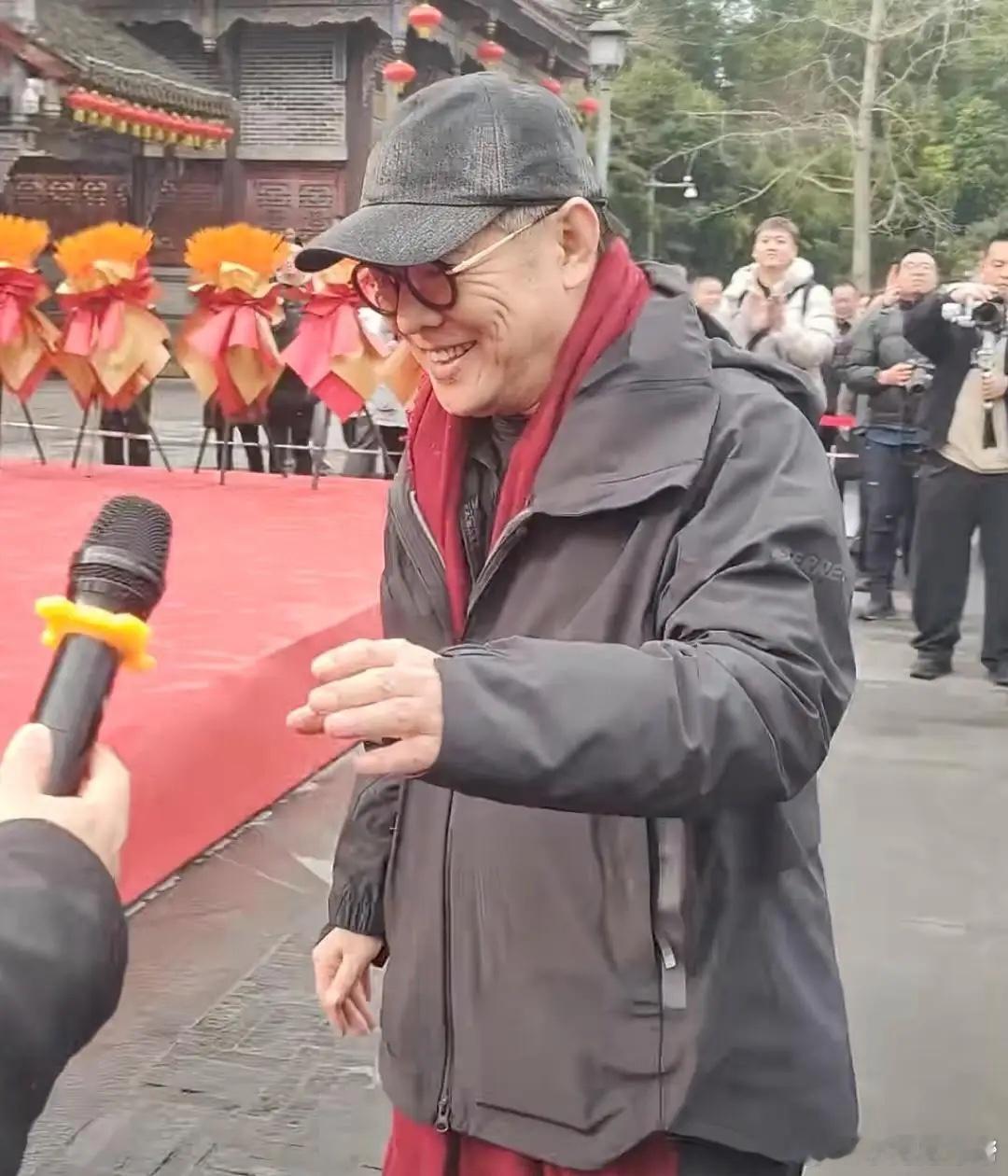 李连杰现身重庆参加短剧开机仪式，他身穿黑色登山服，系着他最爱的红色围巾，头戴鸭舌