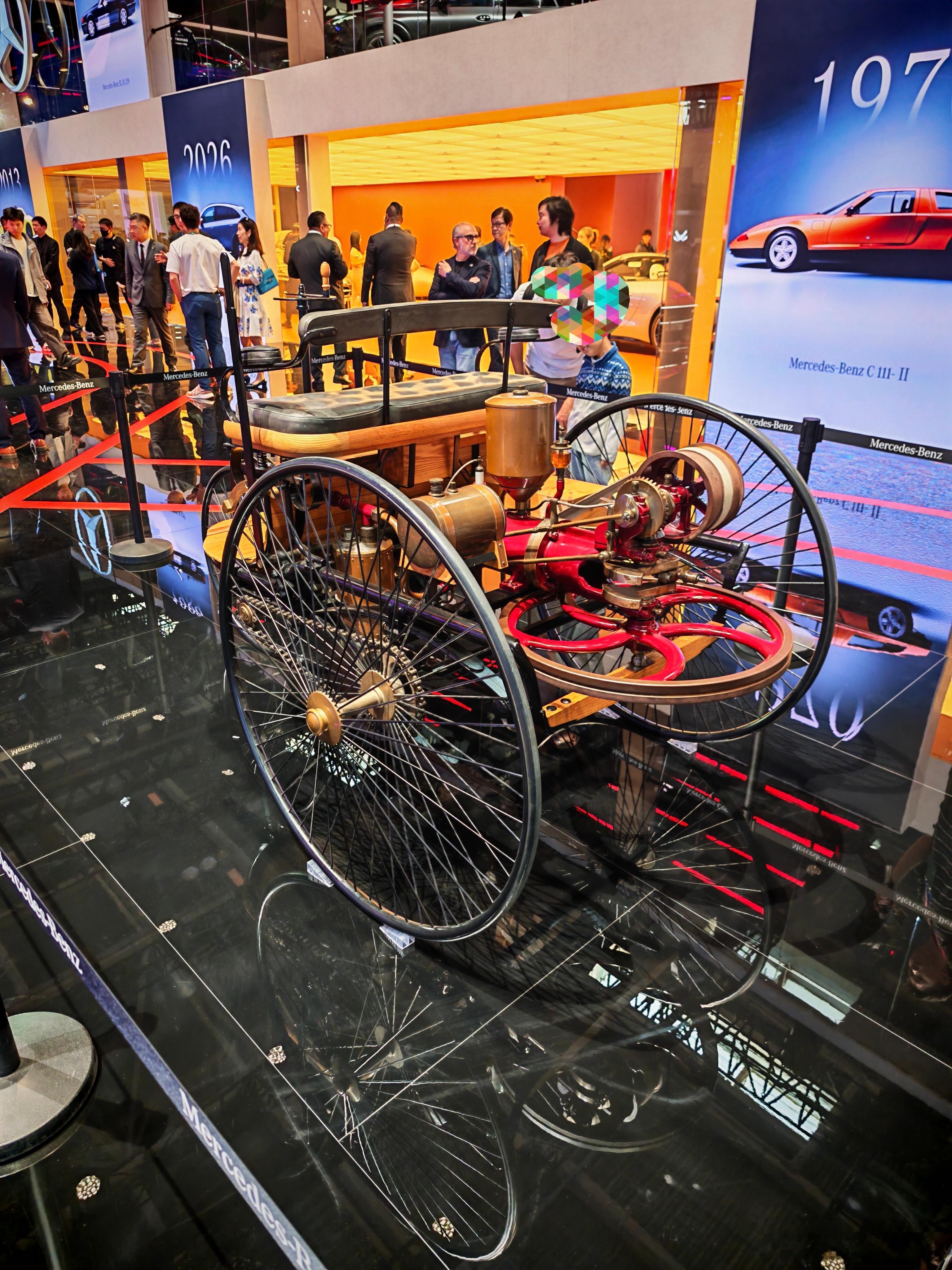 在2026年喧闹的汽车展上，一辆三轮奔驰车静静陈列，红色机械部件如跳动的心脏，诉