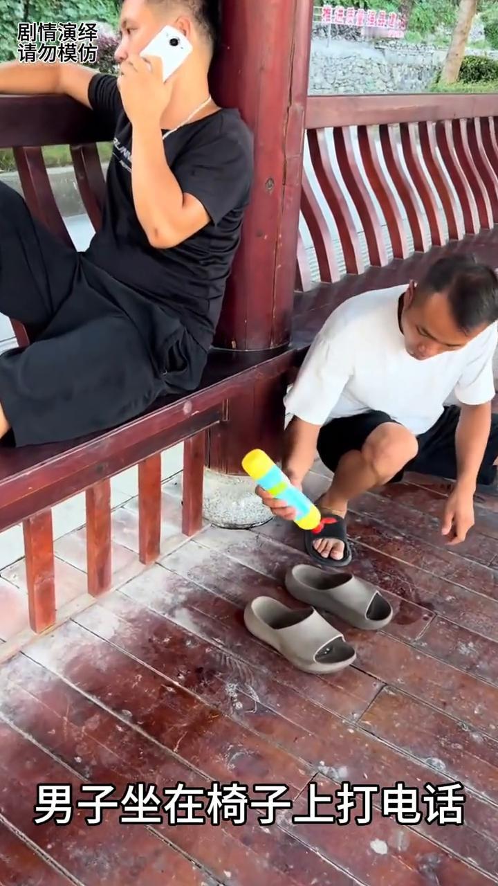 男子坐在椅子上打电话，这时朋友悄悄拿着洗洁精，然后他把洗洁精呲在男子的拖鞋里，随