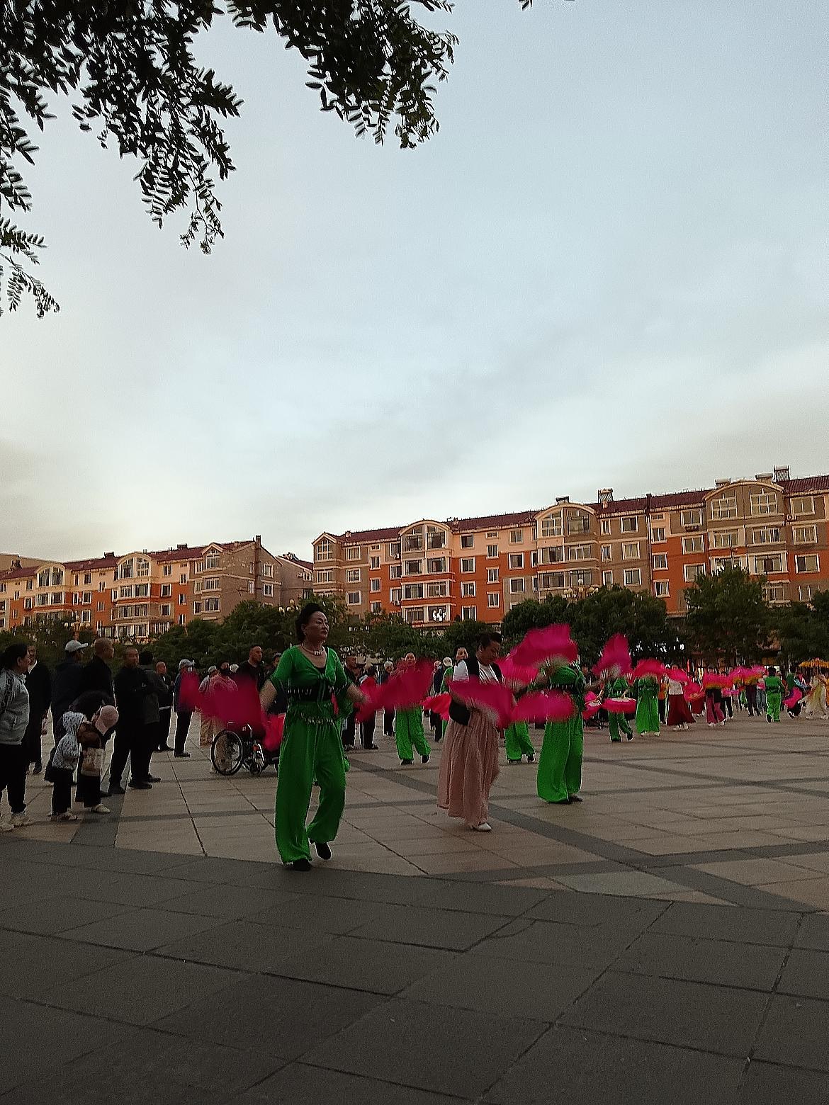 广场上热闹的舞蹈，绿衣舞者翩翩起舞，观众们围观看得津津有味。