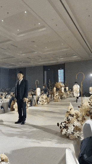 新郎官穿搭黑色西装在婚礼现场等着爱人的出现，此时一位年轻小哥从舞台一边飞奔过来把
