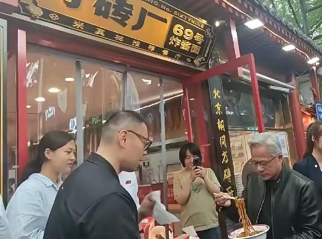 南锣鼓巷今天出了个大新闻——黄仁勋穿着黑皮夹克，站在30度的太阳底下，捧着一碗炸