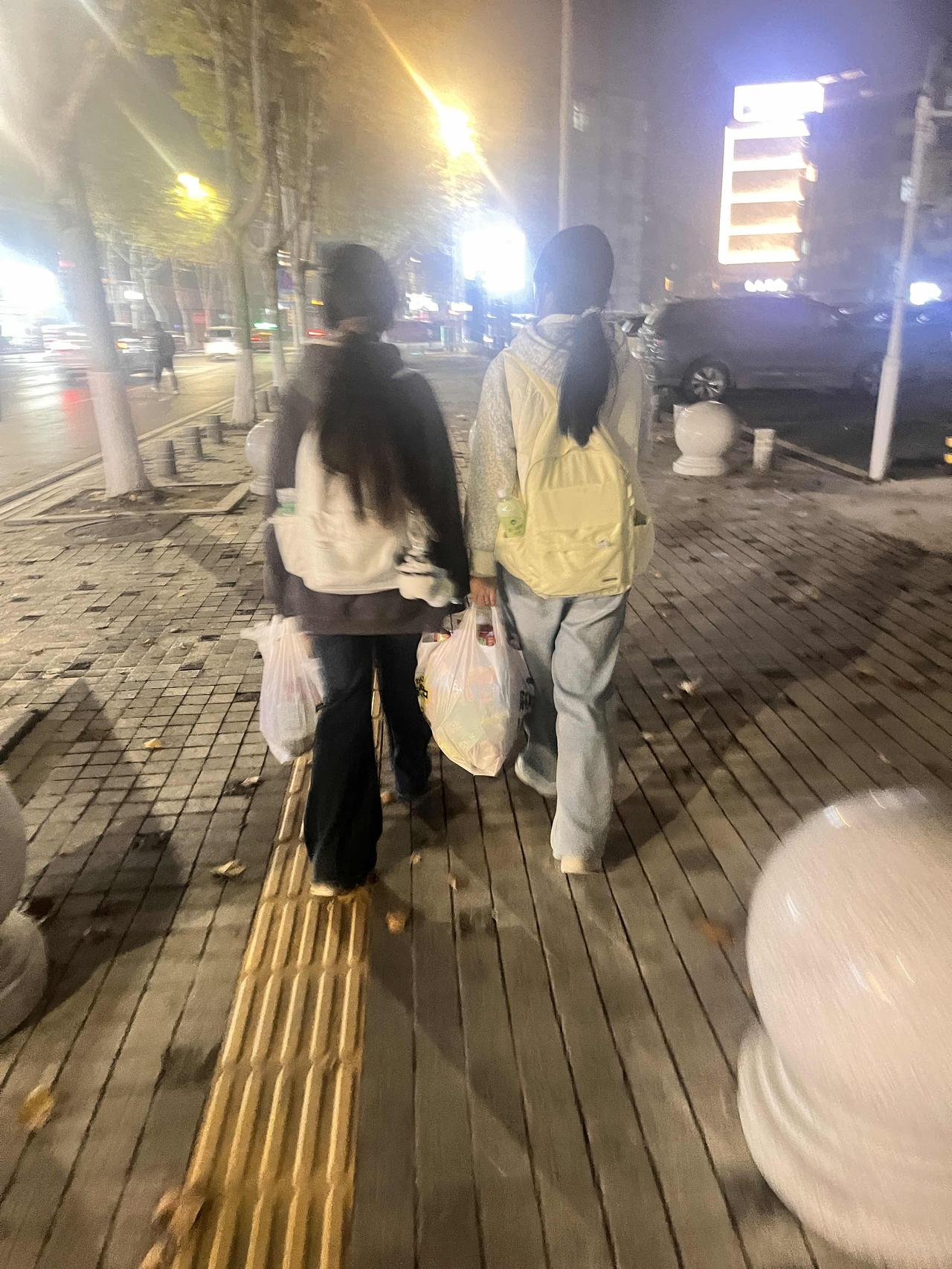 夜晚的街头，路灯把两个女孩的影子拉得长长的。她们并肩走在铺着木质栈道的人行道上，