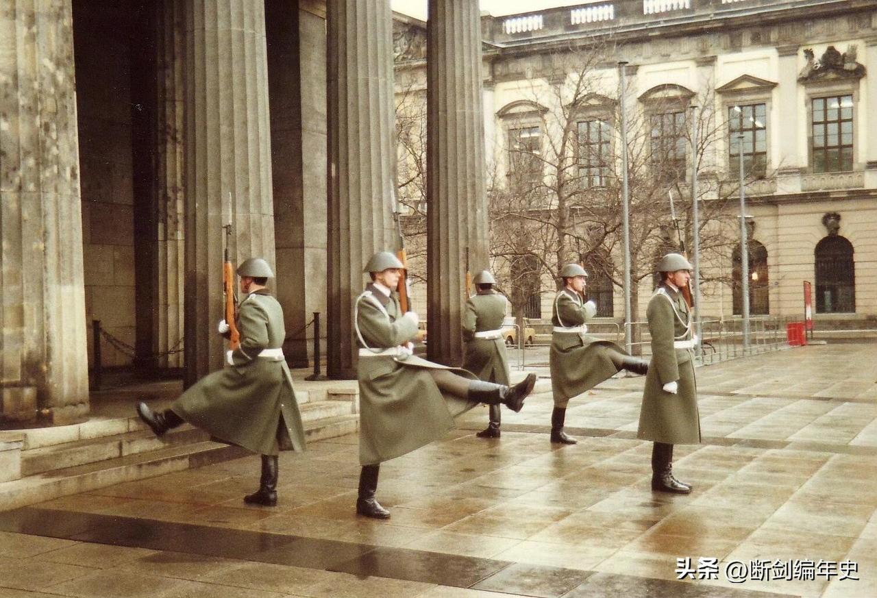 1988年，身着灰绿色长款军大衣、头戴M56钢盔的东德国家人民军士兵，正踢着标志