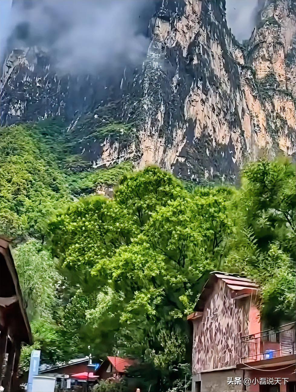 深山实拍，峭壁巍峨，山间云雾缓缓飘荡。满山草木郁郁葱葱，山脚下几座小屋静静依偎在