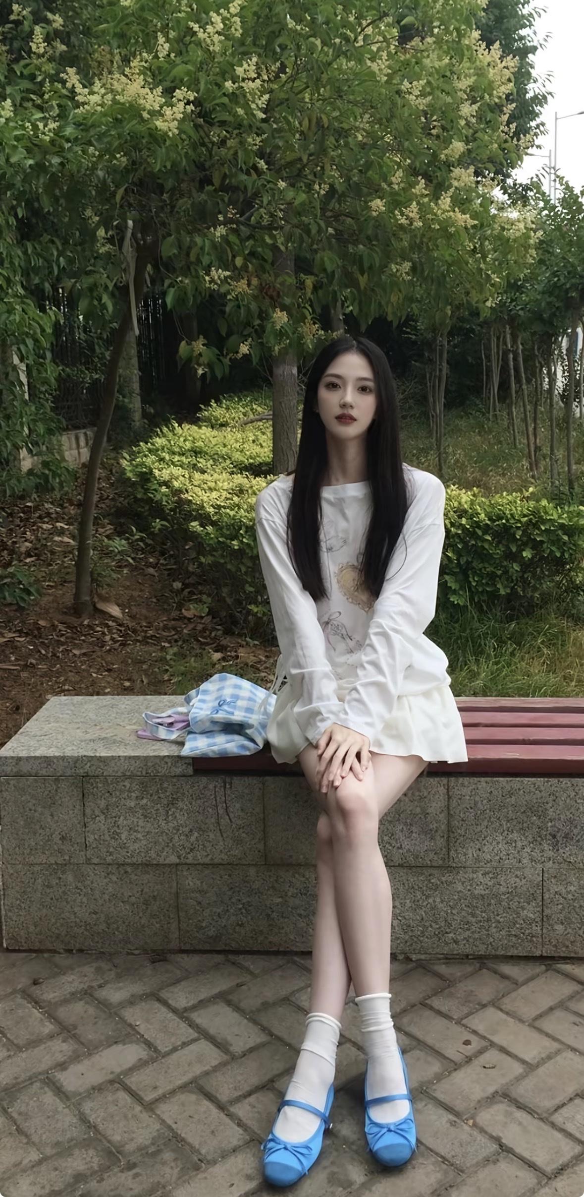 一位黑发长直的女孩坐在绿意盎然的公园石凳上，身穿白色长袖T恤裙，脚踩亮眼的蓝色蝴