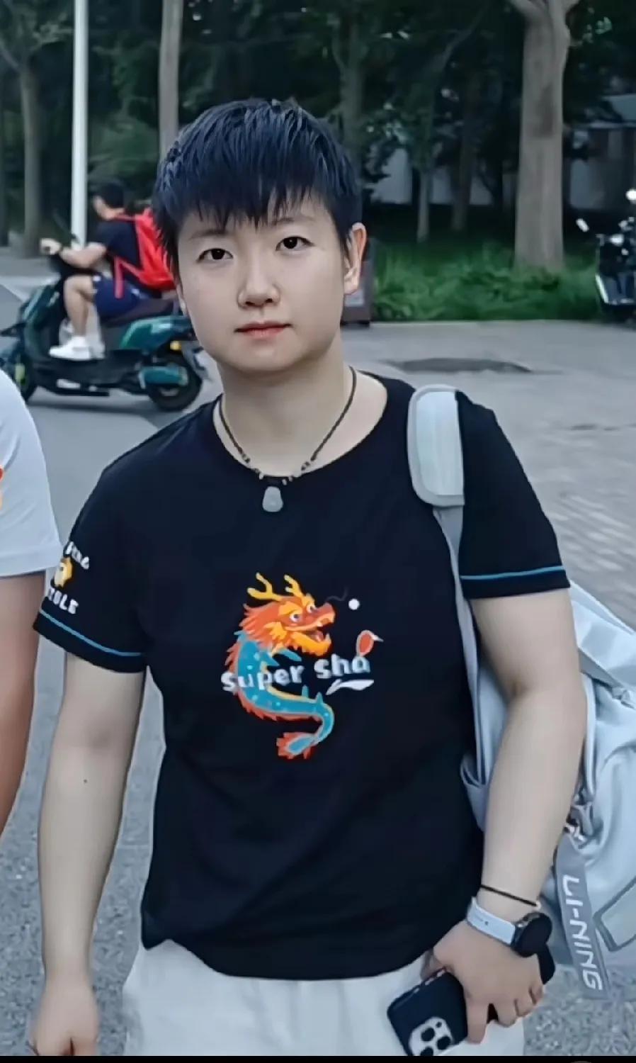 孙颖莎背着单肩包，手里拿着手机，穿黑色印小龙图案的衣服，青春又可爱~ 赛场外的她