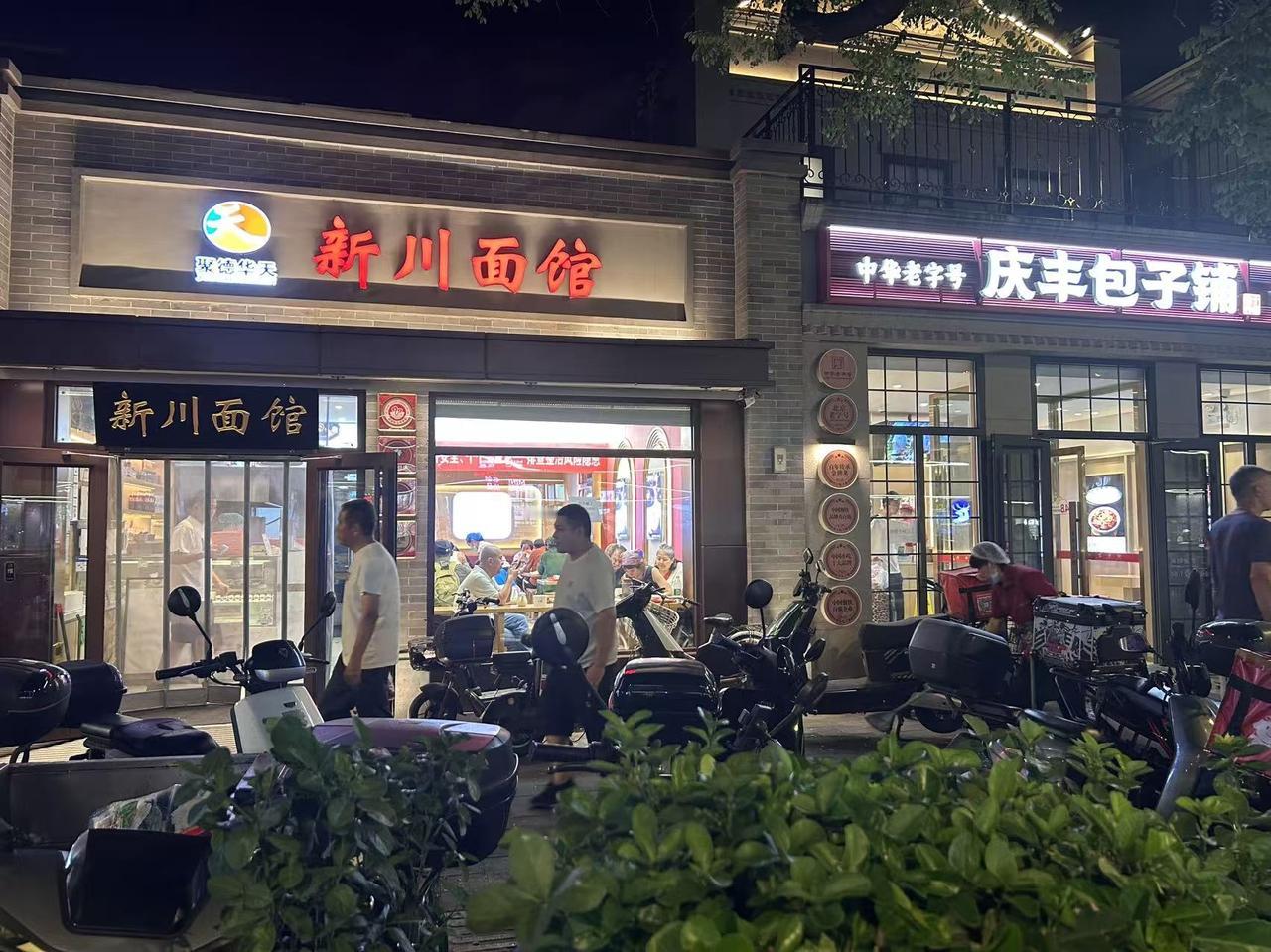 夜色笼罩的新街口，暖光把沿街店铺照亮，烟火气扑面而来。
左边是新川面馆，招牌红色