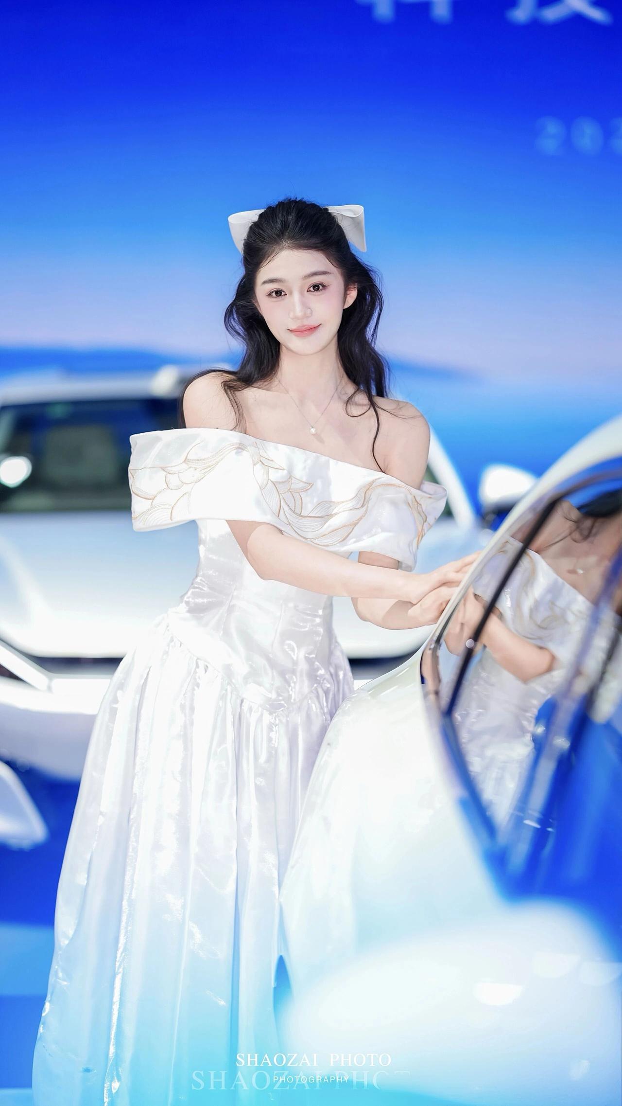 这组照片捕捉了车展上一位气质出众的模特，她身着一袭白色露肩长裙，裙身带有精致的金