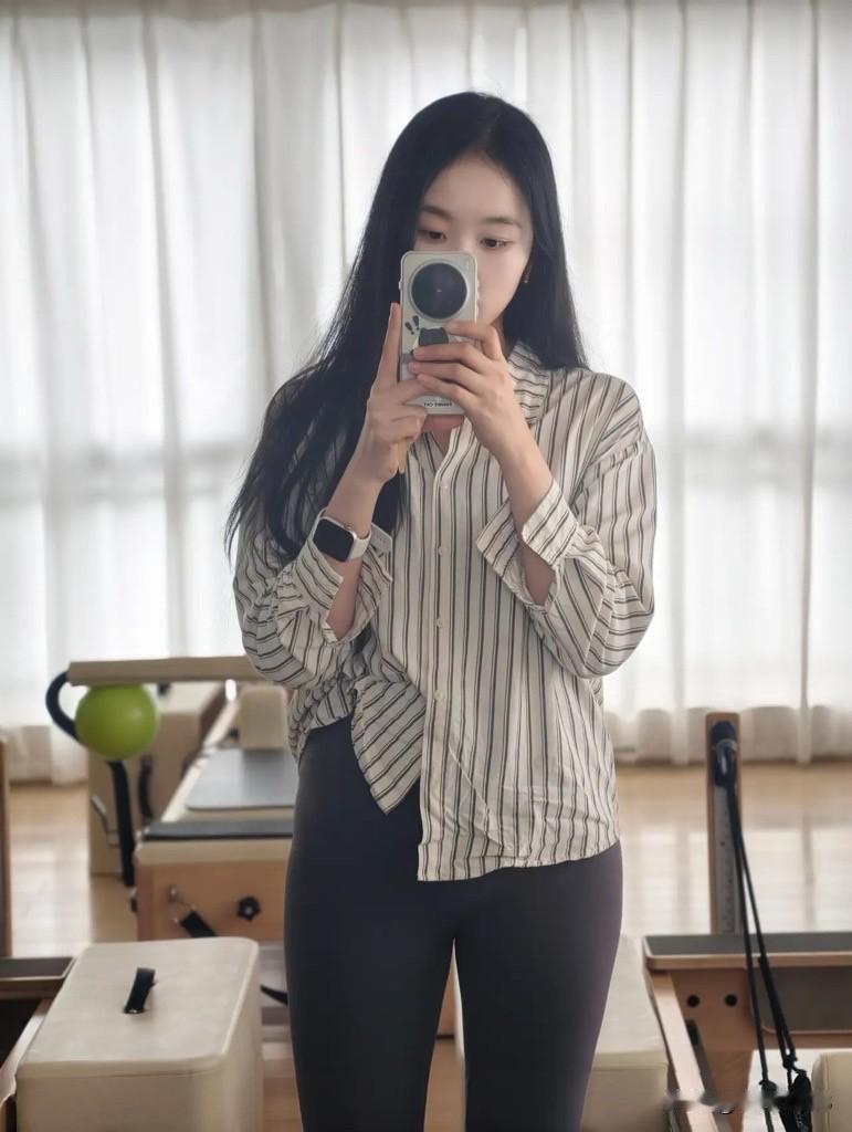 年轻女生长发披肩，上身穿格式衬衣搭配瑜伽修身长裤，双手拿着手机站在室内，柔和的窗