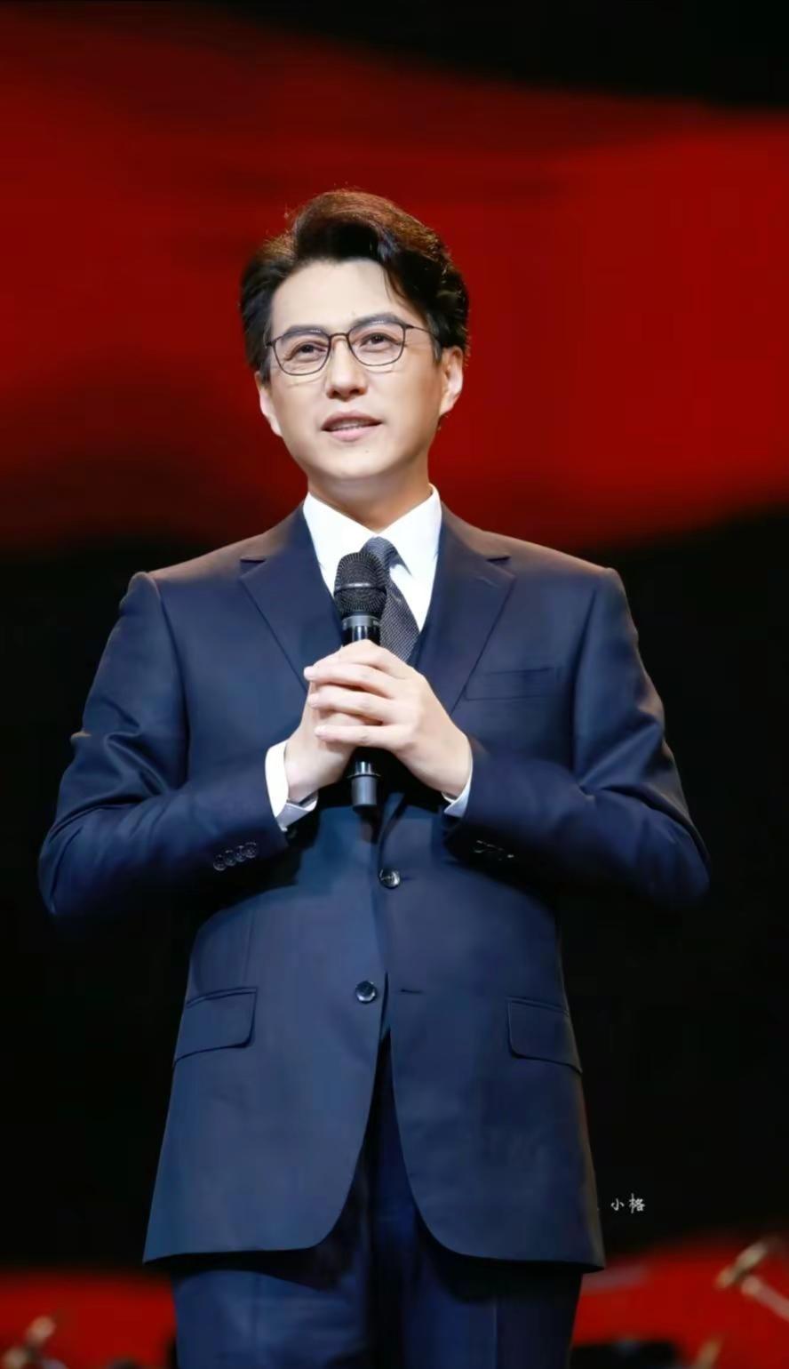 魅力。一袭深色西装沉稳雅致，红幕映衬下身姿挺拔。话筒在手，神情温和从容，言语间尽