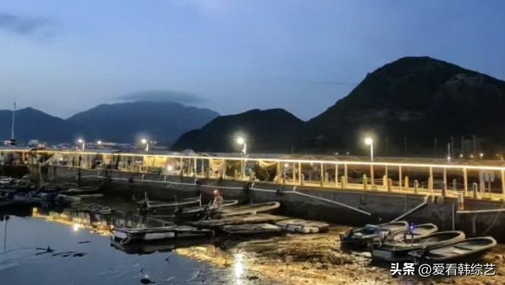 夏日生活打卡季暮色笼罩渔港，远山晕成黛色，堤岸灯带暖光映在静水上。小船静泊滩涂，