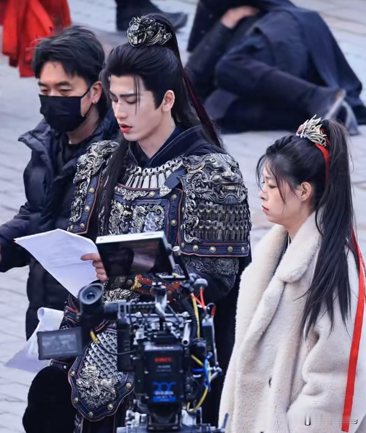 看《逐玉》路透，张凌赫穿着盔甲，现场拿着两张A4纸在镜头前背词，身边站着田曦薇替
