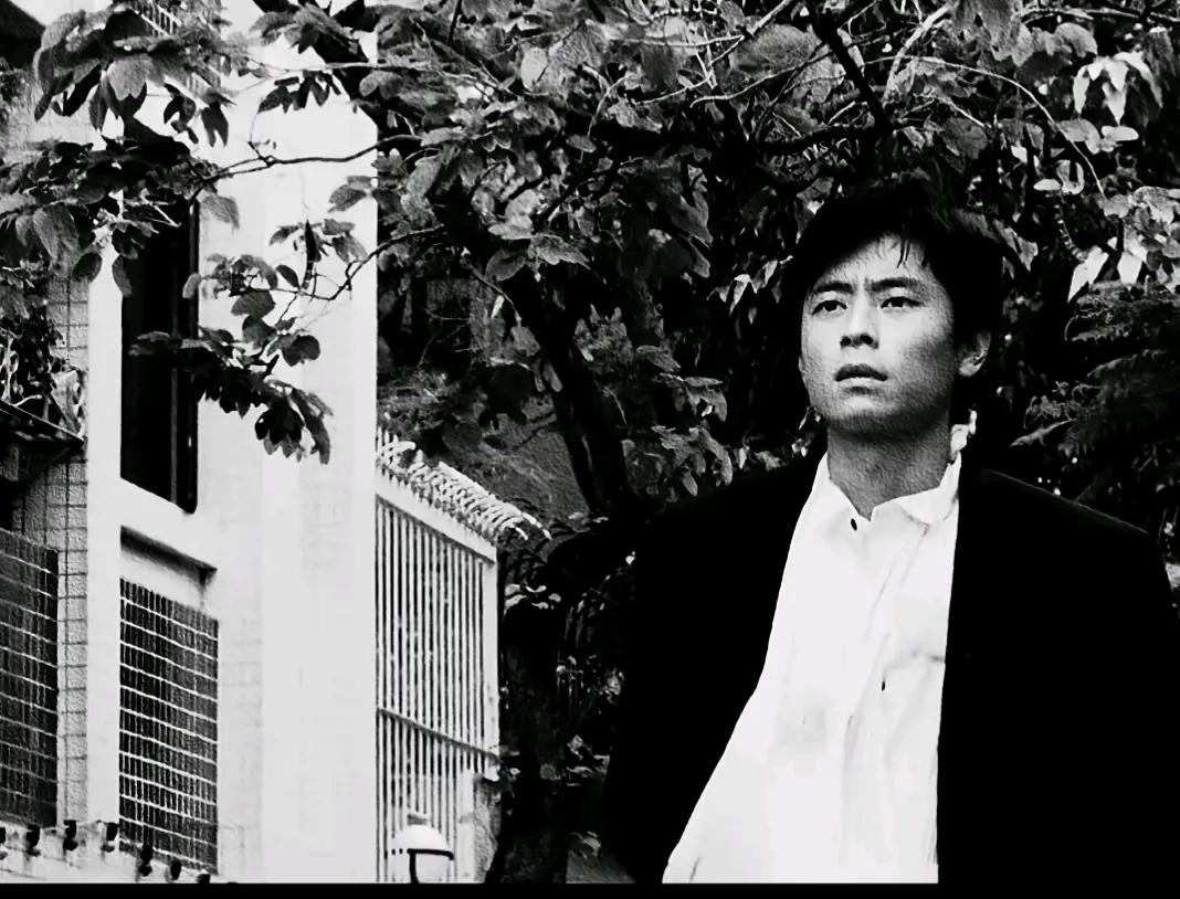 一个男人站在树旁，表情严肃。大家帮忙看看这是哪位？看着很面熟，那时候很年轻。