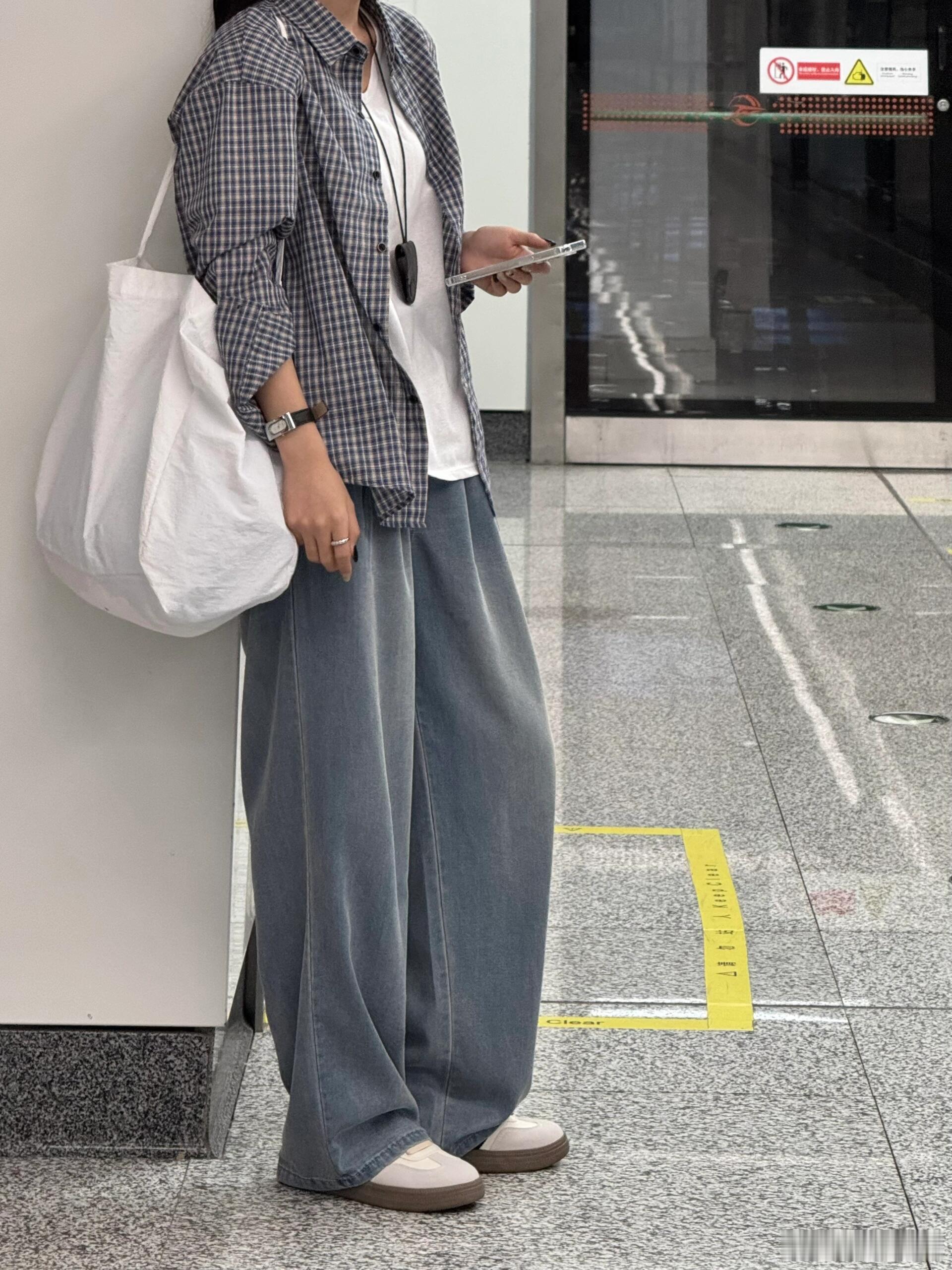 图中女子身着休闲装，站在地铁站内。她上身穿着一件黑白格子衬衫，内搭白色背心，下身