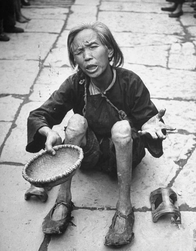 照片中，一位乞讨的老人枯坐在石板路上，身着破旧的深色衣衫，手持空荡的竹编乞讨篮，