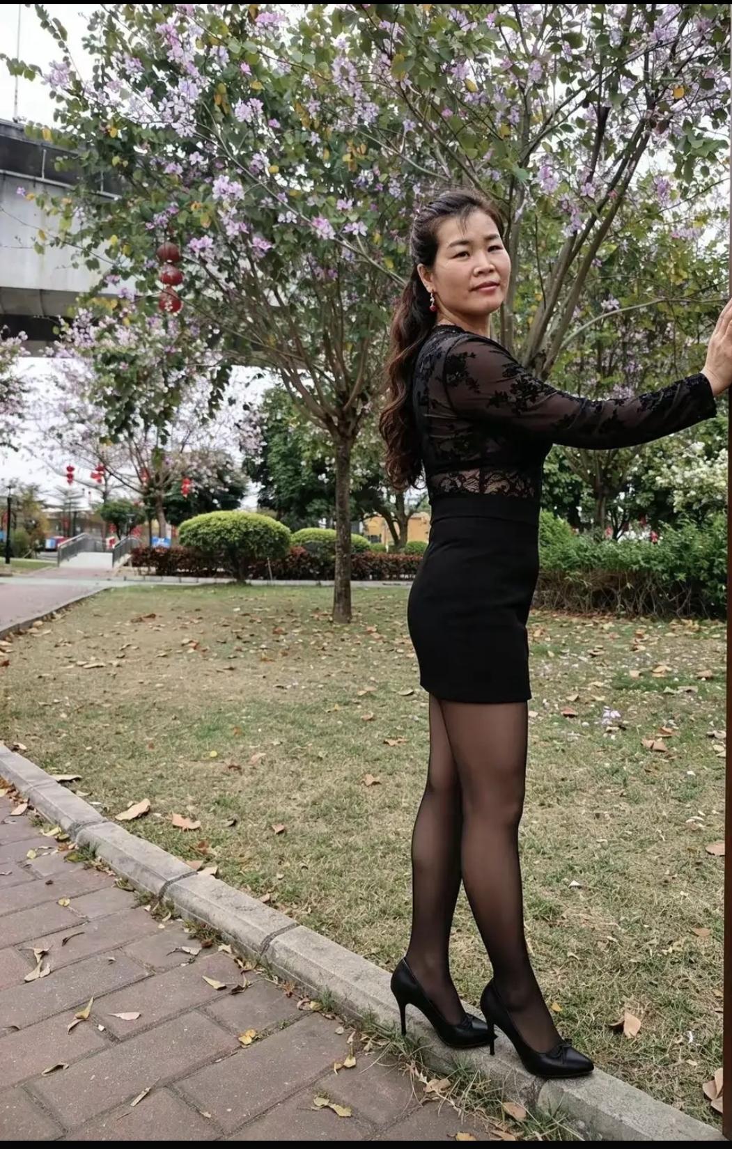 侧身回眸，一手轻扶树干，姿态自然不造作，穿搭精致，黑色蕾丝连衣裙搭配丝袜高跟鞋，