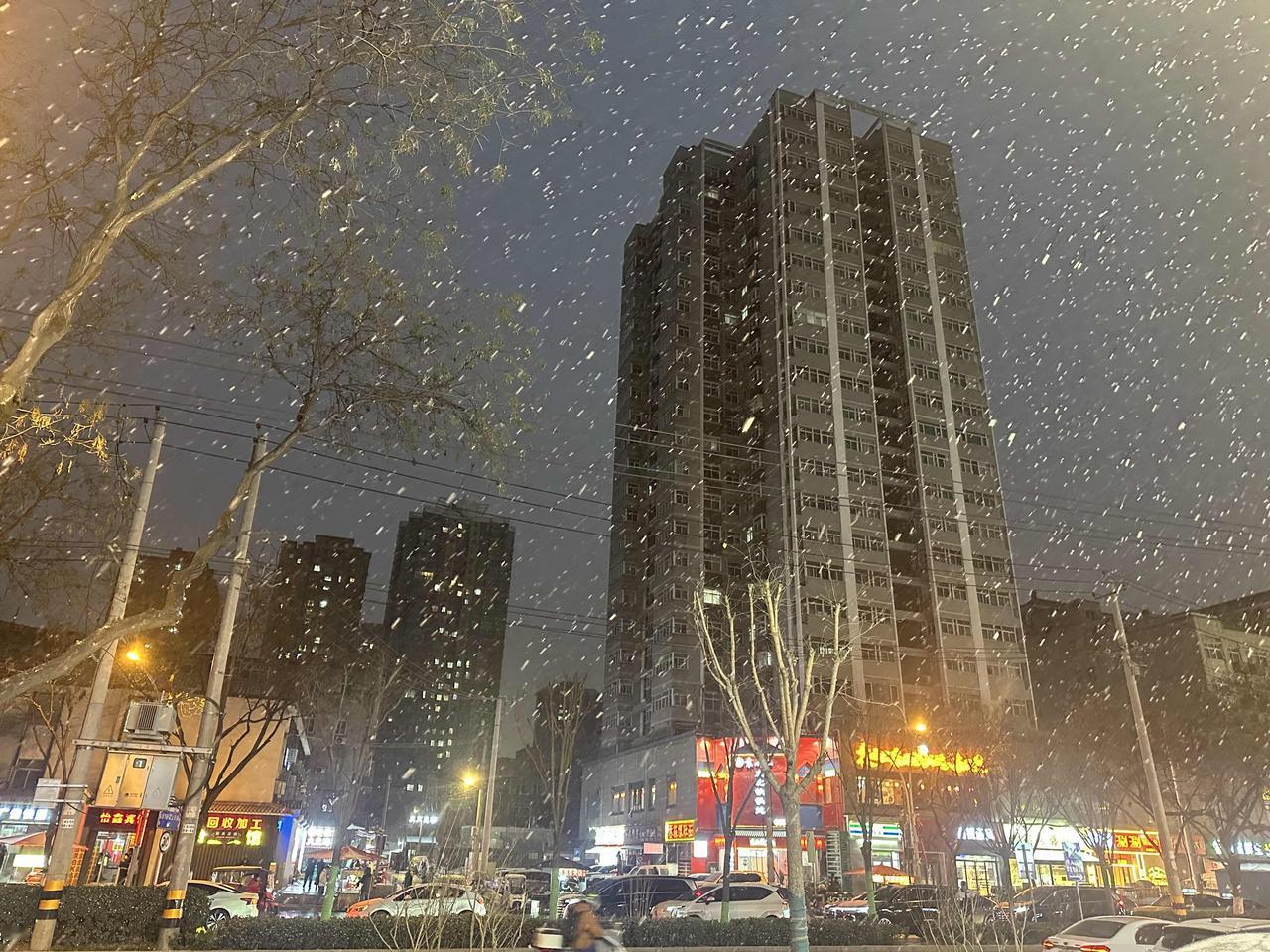 城市的傍晚，雪花飘飘，街上车水马龙，人来人往，街边店铺灯火通明，一派夜晚城市生活