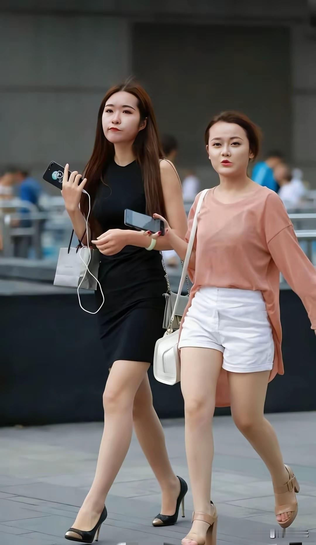 左侧女生一袭黑色紧身无袖连衣裙，搭配浅灰手提包与黑尖高跟鞋，气场十足。右侧女生粉