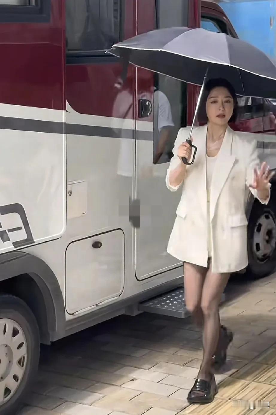 秦岚身穿白色西装套装搭配黑皮鞋，手持雨伞，从车辆旁走过，气场干练