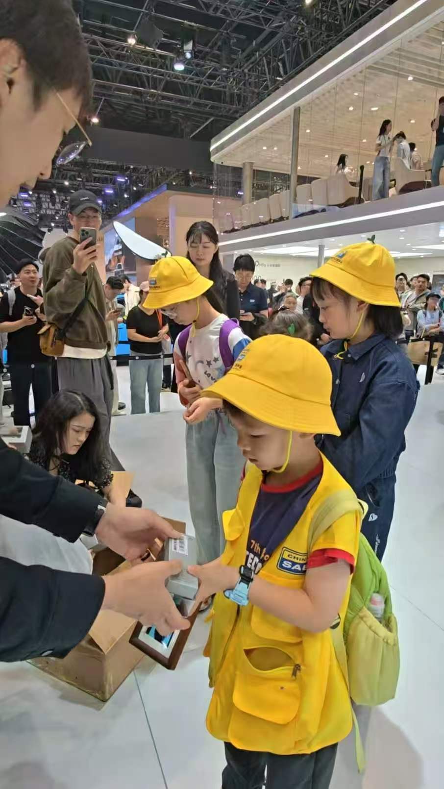 小米展位成小学生研学现场车展小米展台，一群戴小黄帽的小学生把展车围得严严实实，看