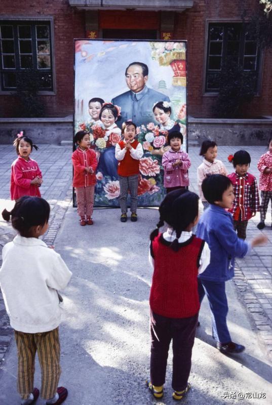 上世纪80年代初，幼儿园小朋友们围成一圈，拍着手又唱又跳。
在她们身后，立着一幅