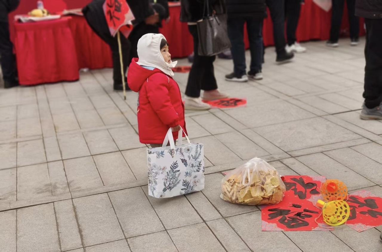 《童趣年味》

红衣小童立街前，
手拎花袋眼儿圆。
福字铺地灯笼俏，
脆片堆旁盼