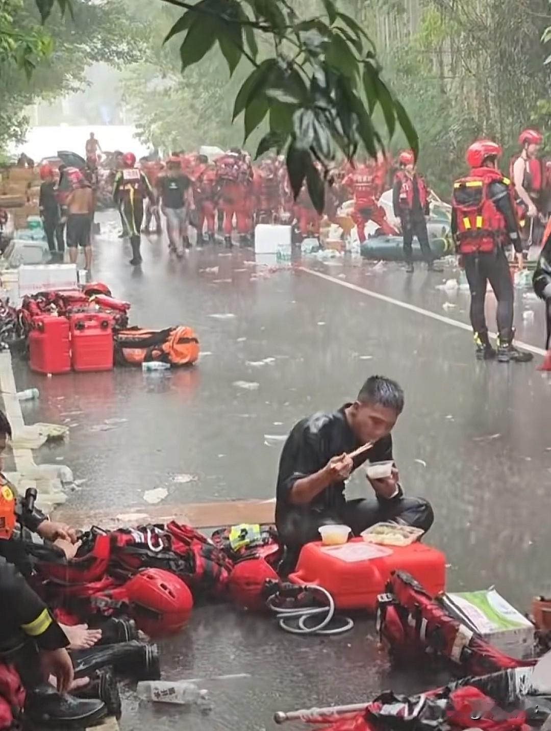 泪目了。一名消防救援人员盘腿坐在路面上就餐，大雨毫无征兆再度倾盆而下，密密麻麻的